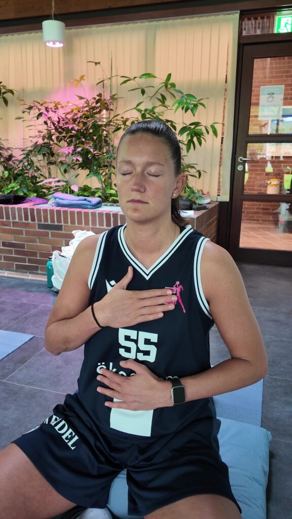 Une femme en chemise de sport noire est assise à l’intérieur, les yeux fermés, la main posée sur sa poitrine et son ventre ; Elle semble calme et concentrée – elle pratique peut-être la respiration consciente ou la méditation, car les bains thermaux optimisent votre entraînement.