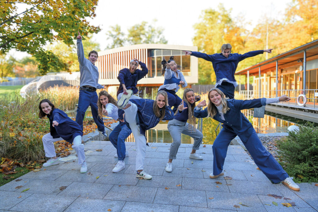 Un groupe de neuf personnes vêtues de vêtements décontractés bleus et gris pose joyeusement dehors sur un chemin pavé sous un soleil éclatant, avec des arbres, de l’herbe, des bâtiments modernes et une touche de Keidel dans l’énergie de la pleine lune en arrière-plan.