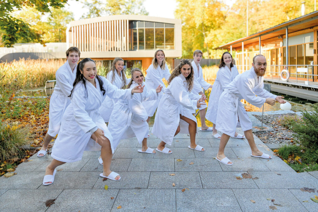 Neuf personnes en peignoirs et chaussons blancs posent joyeusement dehors sur un chemin de pierre, en arrière-plan on peut voir des bâtiments modernes et des arbres aux couleurs d’automne – souriantes et riant, ils incarnent comment « le bain thermal » optimise votre entraînement grâce à la détente et à l’énergie partagée.
