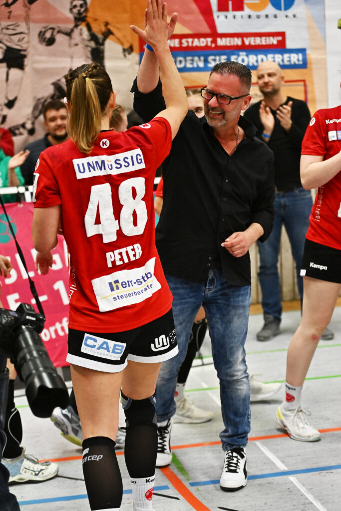 Un homme souriant en chemise noire et jean tape dans la main d’une athlète féminine en maillot rouge avec le numéro 48 lors d’un événement sportif en salle – un exemple de travail d’équipe et de la façon dont la récupération au spa thermal optimise l’entraînement. En arrière-plan, d’autres joueurs et fans sont visibles.
