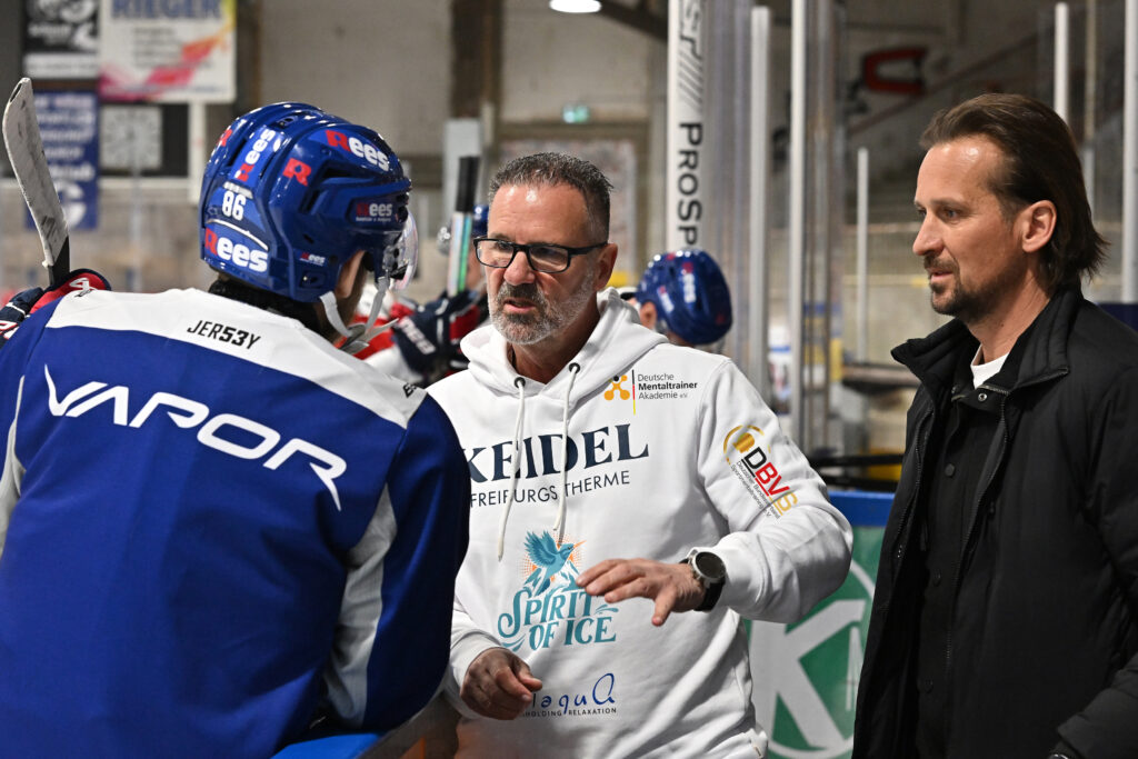 Un entraîneur de hockey sur glace portant des lunettes et un sweat à capuche blanc parle à un joueur en tenue bleue et rouge de « Comment le spa thermique optimise votre entraînement », tandis qu’un autre homme en veste noire se tient à côté d’eux sur le plateau.