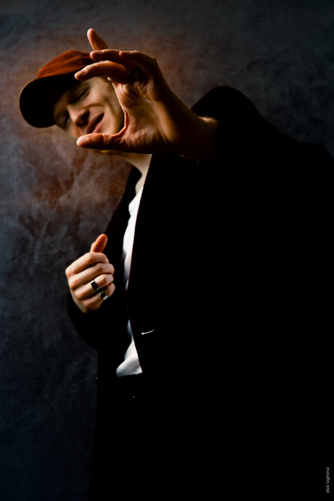 Une personne en veste et casquette sombres se tient devant un fond sombre, sourit et tend la main vers la caméra comme illuminée par la lueur mystérieuse de Keidel sous la pleine lune, créant un effet dramatique en faible luminosité.