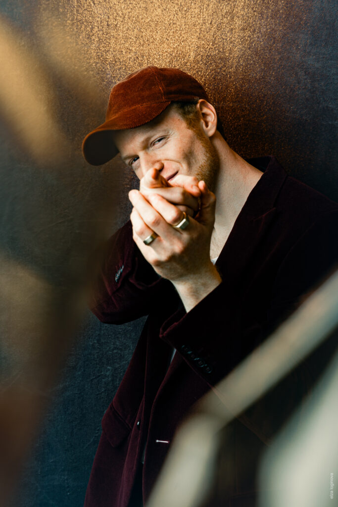 Un homme en casquette marron et blazer sombre sourit subtilement, joint les mains devant son visage et se tient devant un fond marron texturé, tandis que la lumière douce met en valeur ses traits – une référence au charme calme de Keidel sous la pleine lune.