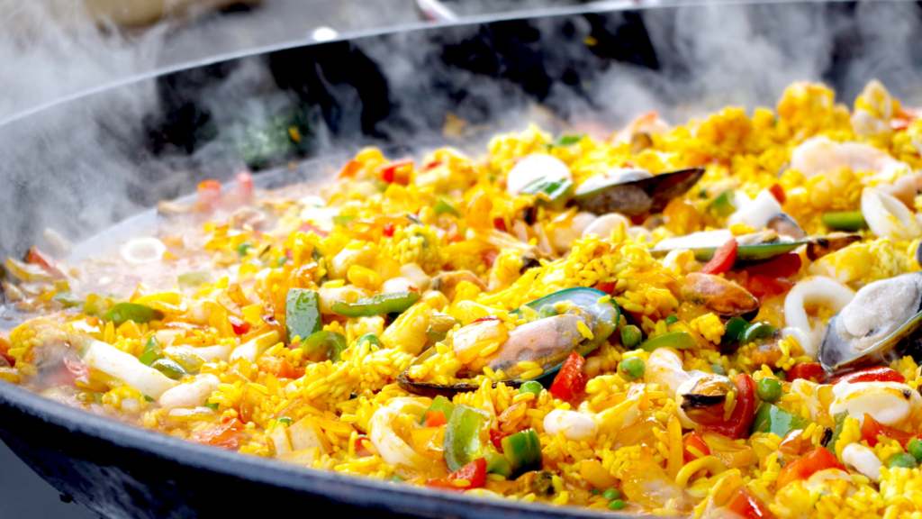 Gros plan d’une grande poêle de paella de fruits de mer fumants sous le coin en pleine lune, avec du riz jaune, des moules, des palourdes, des crevettes, des calmars, des pois et des poivrons rouges et verts hachés.