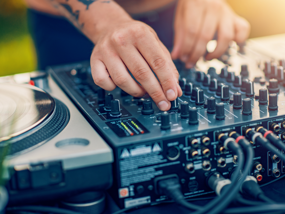 Une personne ajuste les boutons d’un DJ professionnel, une main sur les commandes, l’autre près d’une platine. L’espace extérieur en plein jour capture l’ambiance parfaite de l’été prépayé.