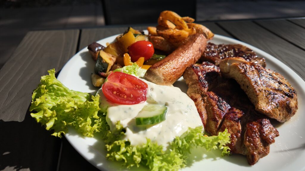 Sur une assiette blanche, on trouve du poulet grillé, du steak, des frites, des légumes frits, de la laitue, une tranche de tomate, des concombres et une sauce blanche crémeuse. Profitez de ce repas vivant en plein air – parfait pour votre prochain repas prépayé d’été.