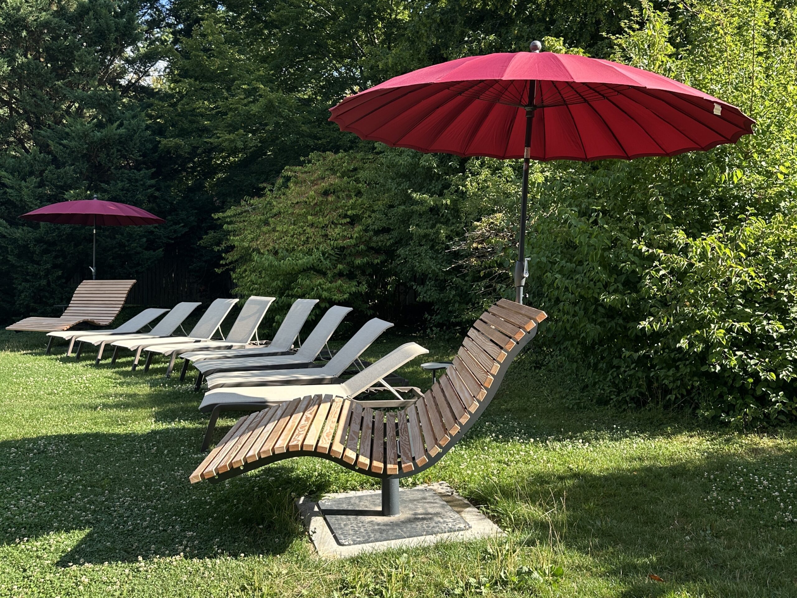 Plusieurs fauteuils en bois avec des parasols rouges sont alignés sur la pelouse, avec en arrière-plan de denses buissons et arbres verdoyants. L’environnement accueillant vous invite à vous détendre en plein air – un endroit idéal pour profiter de votre carte de valeur estivale.