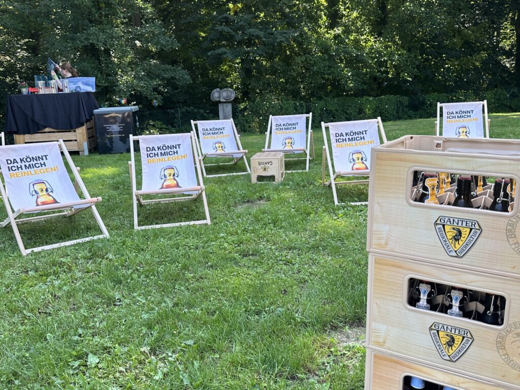 Plusieurs fauteuils lounge en bois avec des panneaux amusants se dressent dans l’herbe à côté de caisses de bière Ganter empilées et d’une carte prépayée d’été, en arrière-plan il y a des arbres et un petit stand à boissons - parfait pour un événement en plein air.