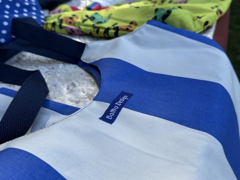 Gros plan d’un sac fourre-tout rayé bleu et blanc avec l’inscription « Bondi Beach », à côté de tissus colorés à motifs et d’une carte prépayée d’été en arrière-plan.