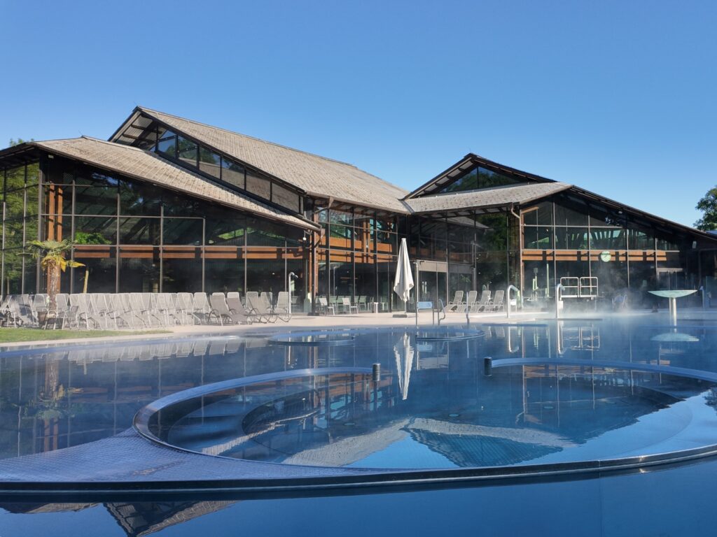 Un bâtiment moderne avec de grandes fenêtres vitrées et des poutres en bois est situé derrière une piscine extérieure fumante, où les invités peuvent se détendre sur des chaises longues sous un ciel bleu — parfait pour se détendre avec la carte prépayée d’été.