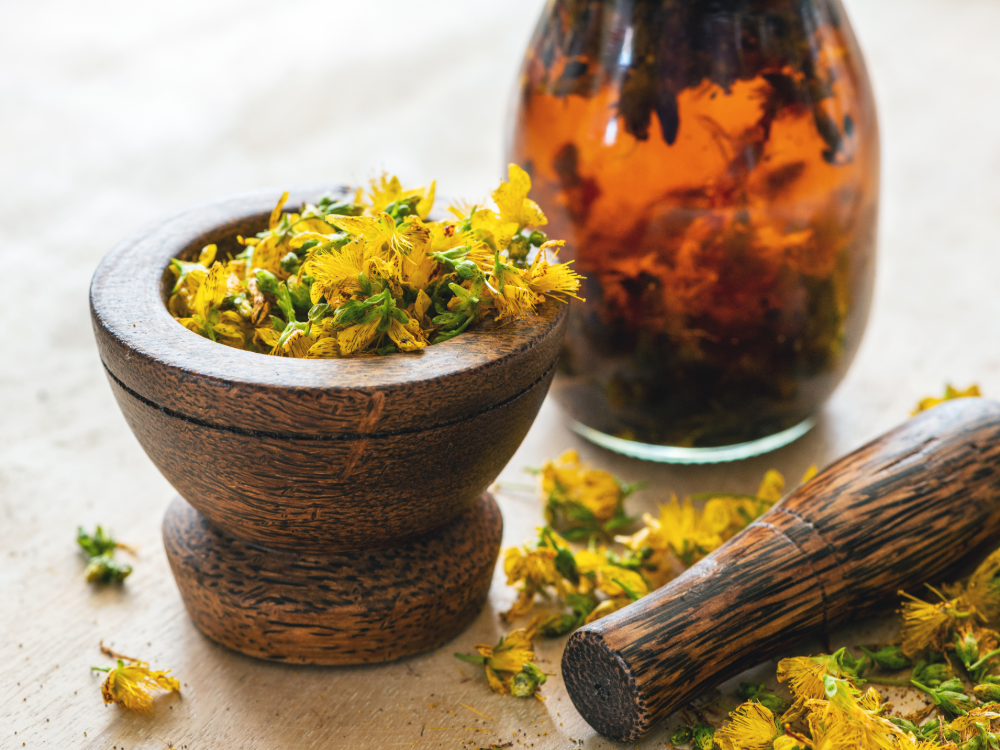 Un mortier en bois rempli de fleurs jaunes séchées est posé à côté d’un pilon assorti et d’un pot en verre rempli de liquide ambré et d’herbes flottantes, le tout sur une surface lumineuse qui dégage le charme naturel d’une carte de valeur estivale.