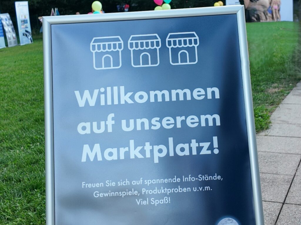 Un panneau sur l’herbe avec des icônes d’étals de marché indique : Bienvenue sur notre marché ! Attendez-vous à des stands d’information passionnants, des concours, des cartes de valeur estivales, des échantillons de produits et bien plus encore. Des ballons sont visibles en arrière-plan.