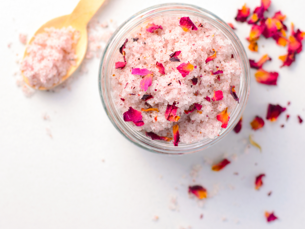 Sur une surface blanche se trouve un pot en verre avec des sels de bain roses mélangés à des pétales de rose séchés, ainsi qu’une cuillère en bois et des pétales éparpillés – le soin de soi parfait après avoir profité de la carte prépayée d’été.