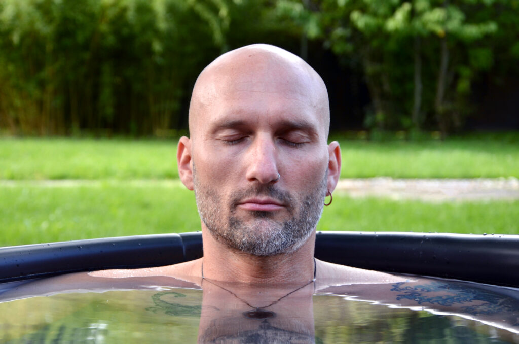 Un homme chauve, barbe et petite boucle d’oreille, est assis les yeux fermés dans une piscine extérieure et se détend. Il s’embrasse dans la glace, se baignant dans les Keidel Thermes et trouve la paix tandis que l’herbe verte et les arbres se brouillent doucement en arrière-plan. 