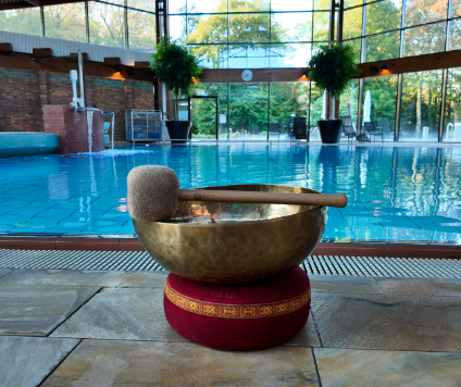 Un bol chantant avec un maillet repose sur un coussin à côté d’une piscine intérieure, entouré de grandes fenêtres, de verdure et de lumière naturelle – un cadre paisible rappelant un rituel « Keidel sous la pleine lune ».